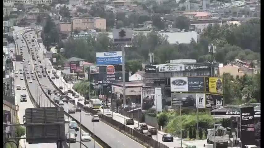 A multi-lane highway with heavy traffic flows through a city with numerous buildings and billboards, on a partly cloudy day.