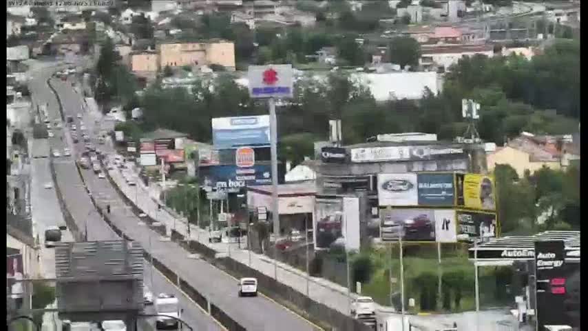 A highway with moderate traffic flows through a city on an overcast day, lined with buildings and billboards.