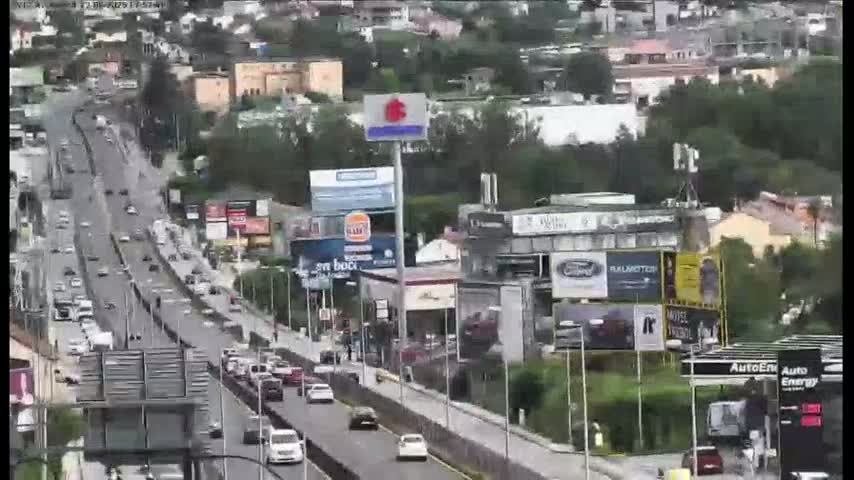 A multi-lane highway with moderate traffic flows through a city on an overcast day, passing by various buildings and billboards.