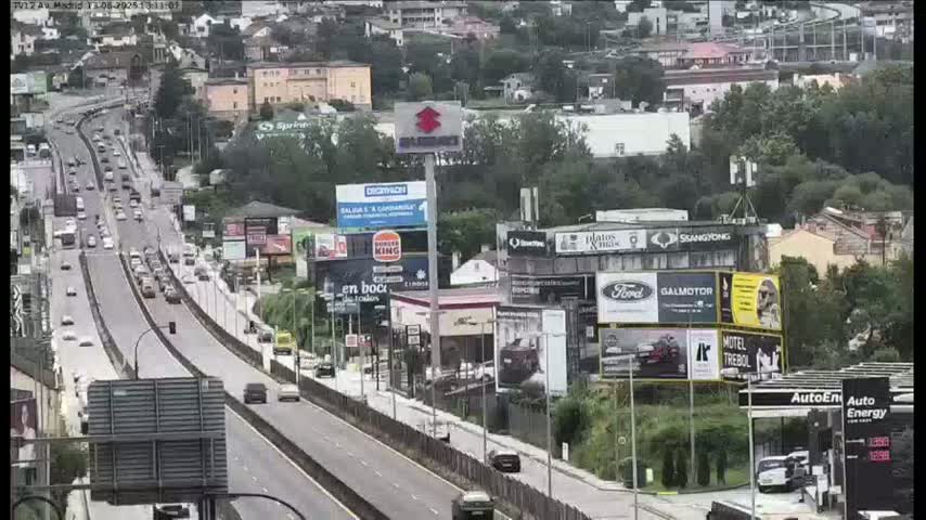 A multi-lane highway with moderate traffic flows through a city under an overcast sky, alongside buildings displaying numerous billboards.