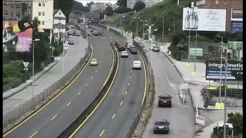 A multi-lane highway with several cars driving in both directions, flanked by buildings and greenery on a sunny day.