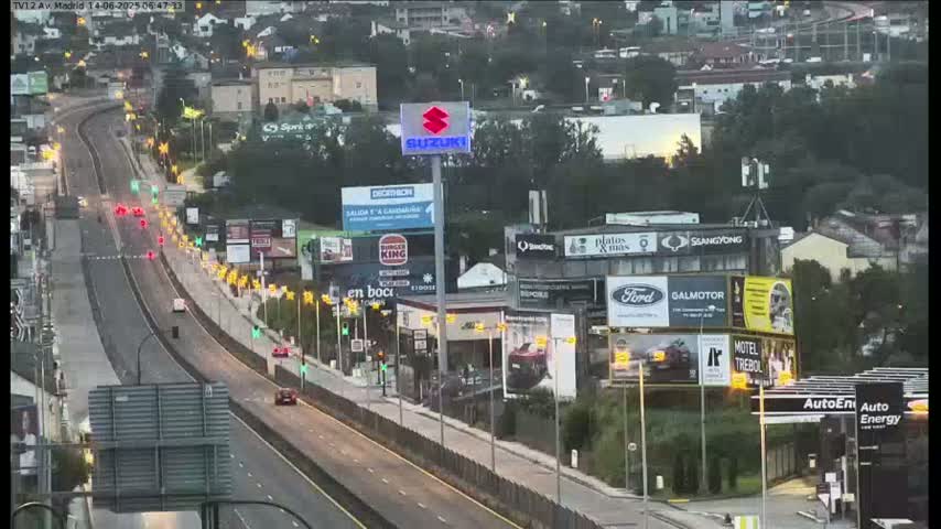 A multi-lane highway with several cars driving in both directions, flanked by buildings and greenery on a sunny day.