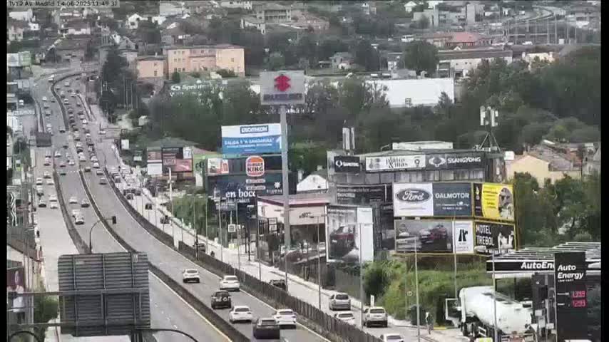 A multi-lane highway with moderate traffic flows through a city with numerous billboards and buildings, under a partly cloudy sky.