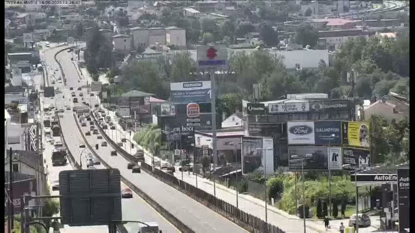 A multi-lane highway with moderate traffic flows through a city on a sunny day, passing by buildings with numerous billboards.
