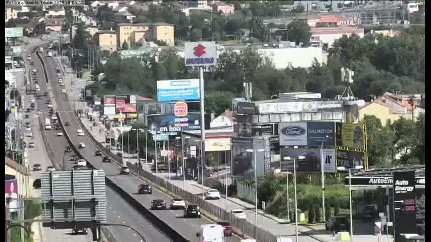 A highway with moderate traffic flows through a suburban area on a sunny day, lined with billboards and businesses.