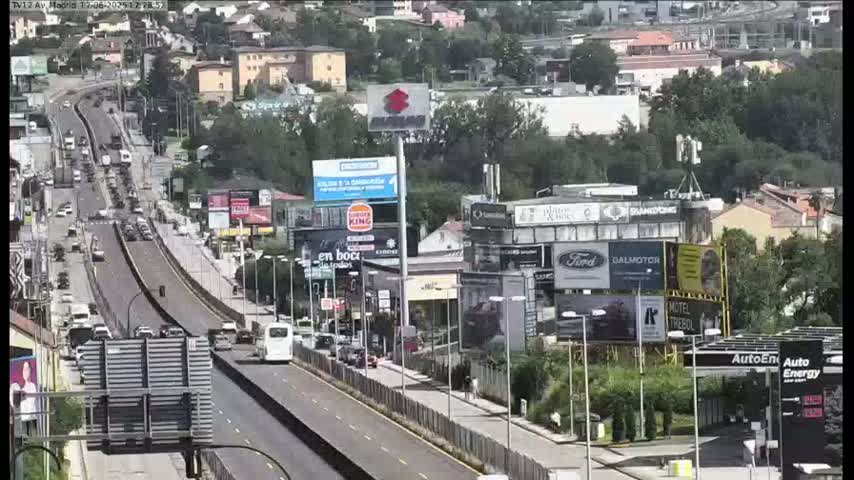 A highway scene on a sunny day shows a multi-lane road with moderate traffic, flanked by buildings with numerous billboards and advertisements.