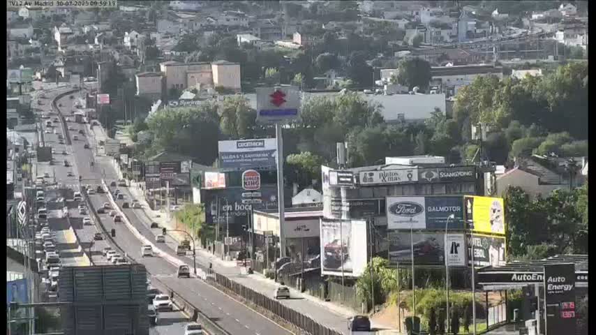 A highway scene on a sunny day shows a multi-lane road with moderate traffic passing by buildings and billboards.