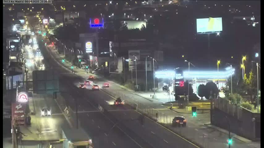 A nighttime, slightly wet roadway with sparse traffic flows past businesses and billboards under streetlights.