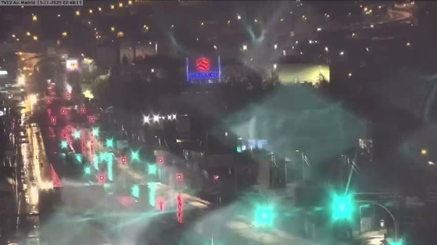An elevated night view captures a multi-lane roadway with the distinct reflections of vehicle lights and streetlights on its wet surface, amidst a foggy or hazy atmosphere.