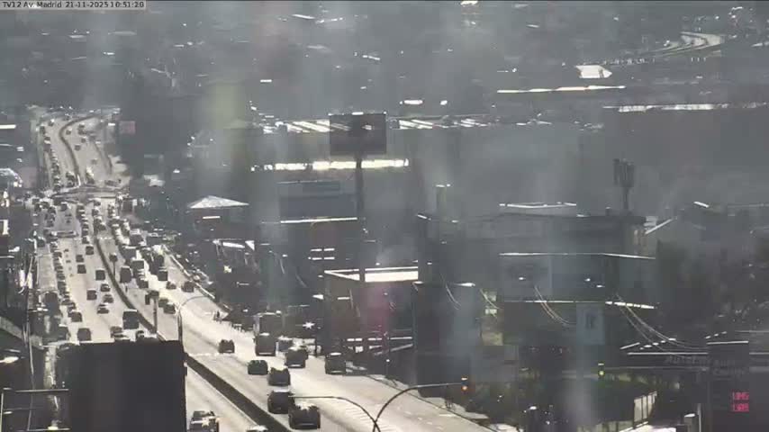 An elevated view shows a multi-lane highway filled with heavy traffic winding through a hazy urban landscape under bright daytime conditions.