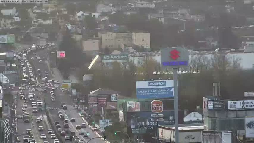 An elevated view captures a busy multi-lane highway congested with vehicles, flanked by numerous buildings and some bare trees, under an overcast sky.