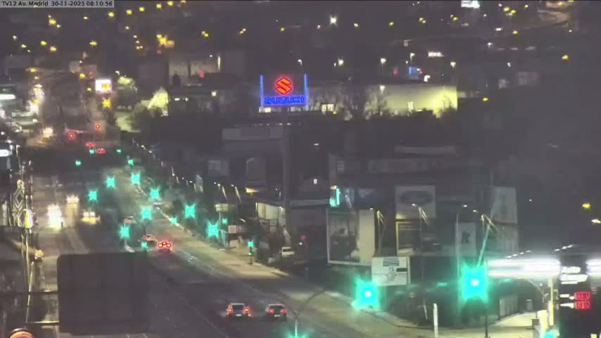 An overhead view captures a bustling city street at night, with vehicles driving on a wet road reflecting bright streetlights and the glowing signs of surrounding buildings, including a prominent blue illuminated sign in the distance.