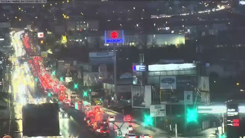 A wet urban highway at night is illuminated by the red taillights and white headlights of heavy traffic, with city lights visible in the background.