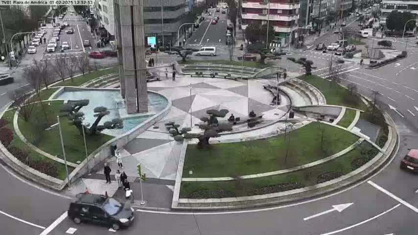 A circular plaza with a central fountain and landscaping, surrounded by a roadway with cars, under an overcast sky.