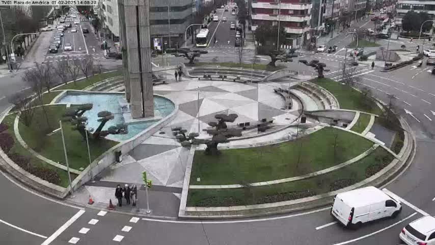 A circular plaza with a fountain and landscaping is surrounded by roads with cars, under an overcast sky.