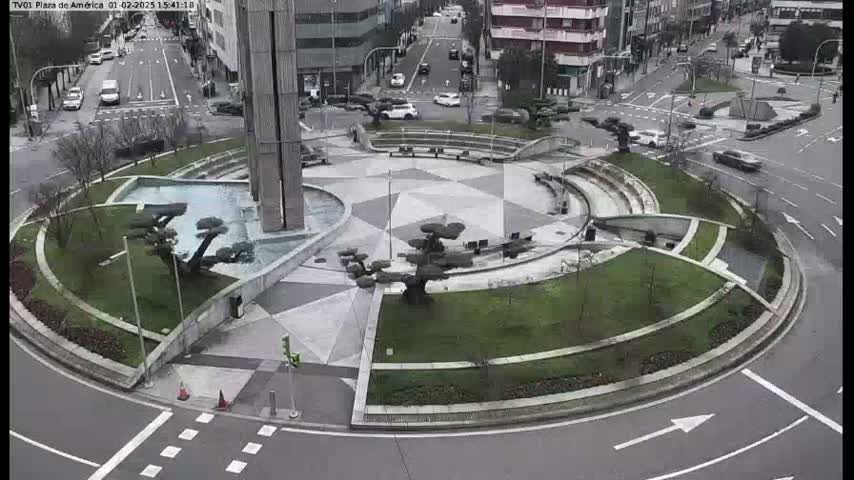 A landscaped roundabout with a central fountain and several trees is situated in the middle of a city intersection, on an overcast day.