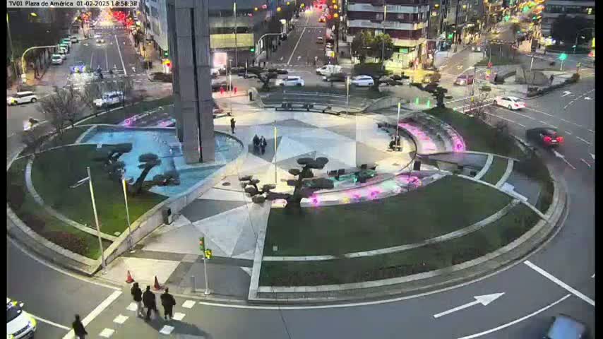 A nighttime aerial view shows a circular plaza with a water feature and several small trees, surrounded by a roadway with cars and pedestrians.