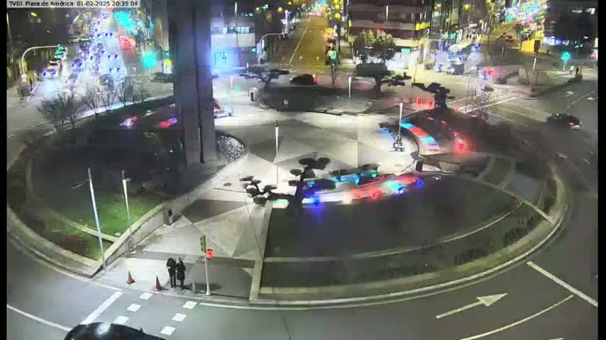 At night, emergency vehicles with flashing lights are in a roundabout surrounded by a plaza with landscaping and parked cars nearby.
