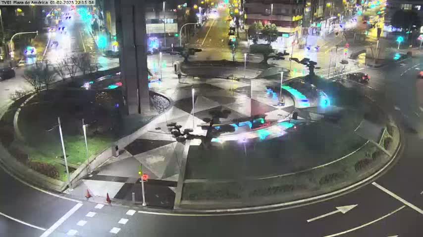 A nighttime aerial view of a roundabout with geometric landscaping and sparse traffic on surrounding roads under wet conditions.