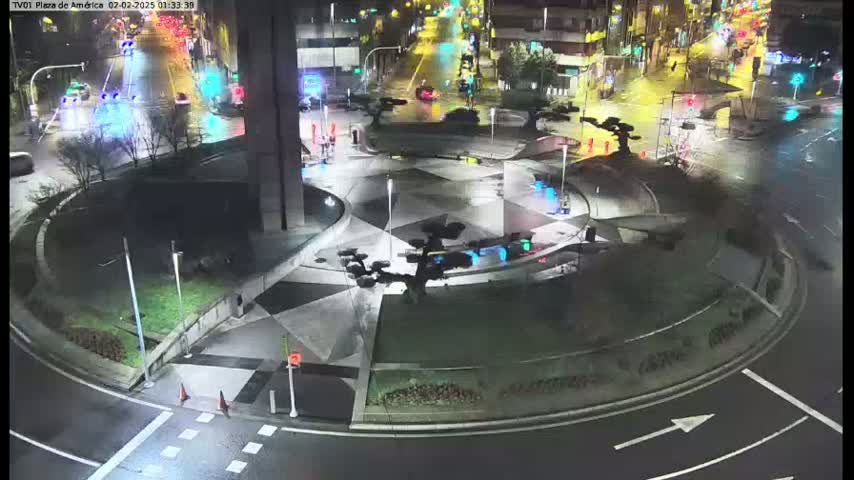 A nighttime, wet, aerial view shows a circular plaza with geometric patterned pavement, sparse landscaping, and several stylized dark sculptures in the center, surrounded by roadways with minimal traffic.