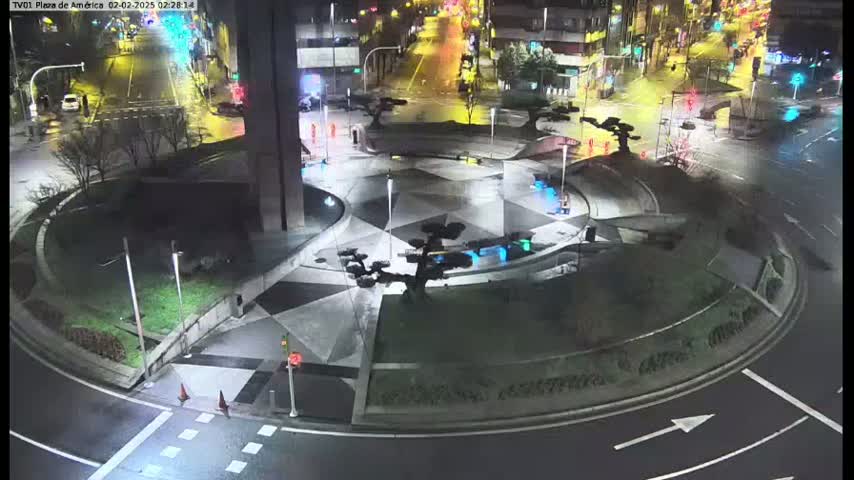 A nighttime aerial view of a circular plaza with geometric paving, small trees, and surrounding roads showing some vehicle traffic under wet conditions.