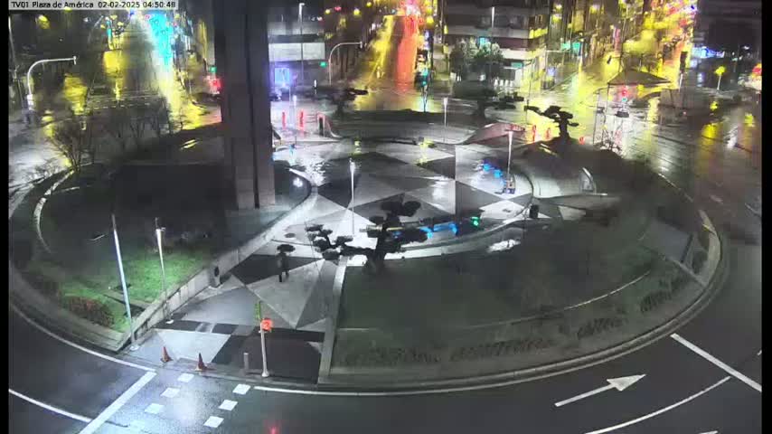 A wet, nighttime scene shows a circular plaza with geometric patterns, small trees, and sparse pedestrian traffic around a roundabout.