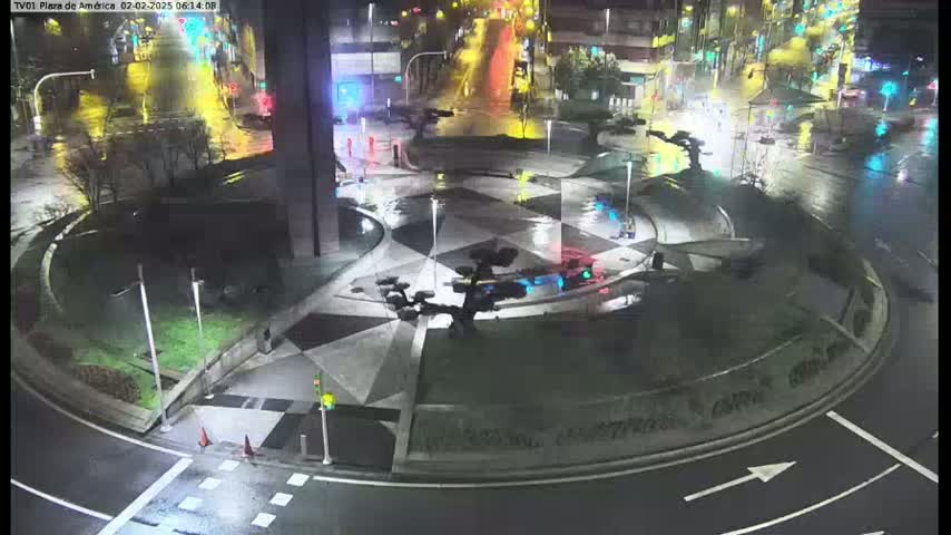 A wet, nighttime scene shows a circular plaza with geometric designs, several dark metal sculptures, and surrounding roadways with minimal traffic.