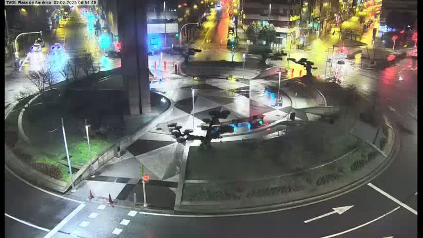 A wet, nighttime scene shows a roundabout with a geometrically patterned center, surrounded by roadways with some vehicle traffic.
