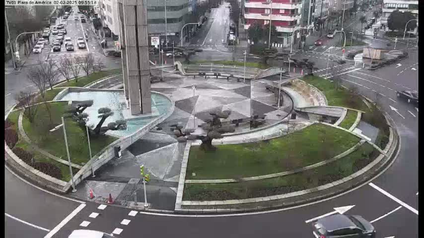 A circular plaza with a fountain and landscaping is situated in the center of a roundabout, with cars traveling around it on a rainy day.