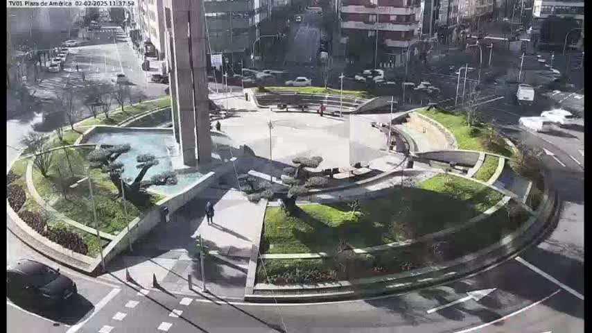 A sunny day reveals a circular plaza with landscaped areas, a central fountain, and surrounding buildings, all encompassed by a roadway with vehicles.