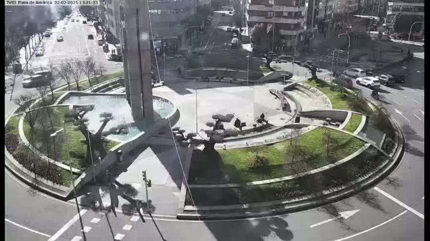 A sunny aerial view of a circular plaza with a fountain, landscaping, and a few people sitting on benches, surrounded by a roadway with cars.