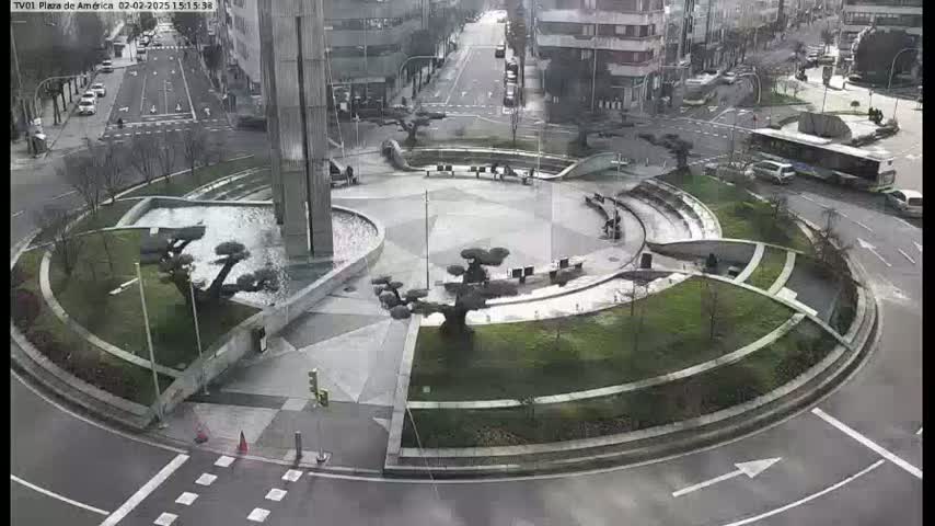 An overcast day shows a circular plaza with a fountain, landscaping, and several small trees, surrounded by roadways and buildings.