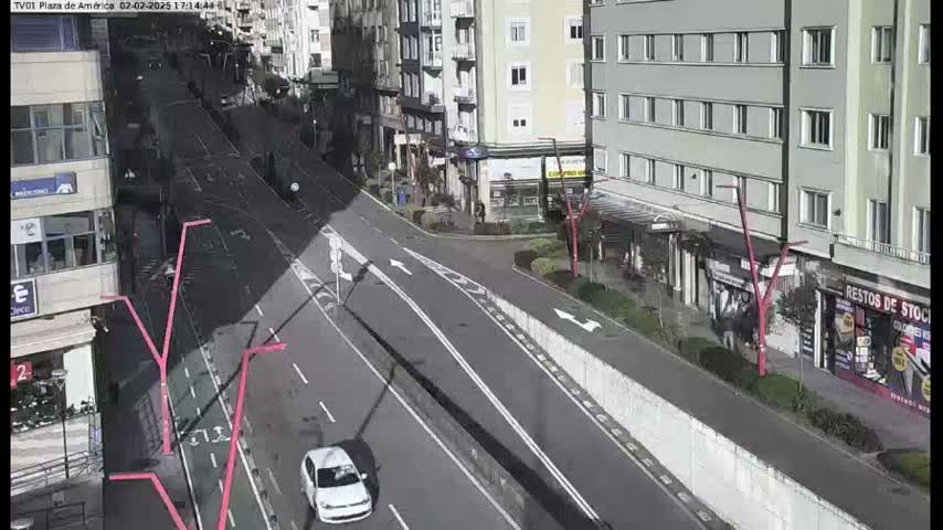 A mostly empty city street with buildings on either side, a white car driving down the road, and a sunny day.