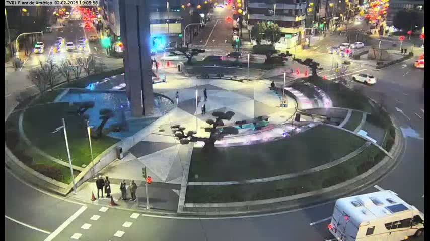 A nighttime aerial view of a circular plaza with a fountain, landscaping, and a few pedestrians, surrounded by city streets with cars and a camper van.