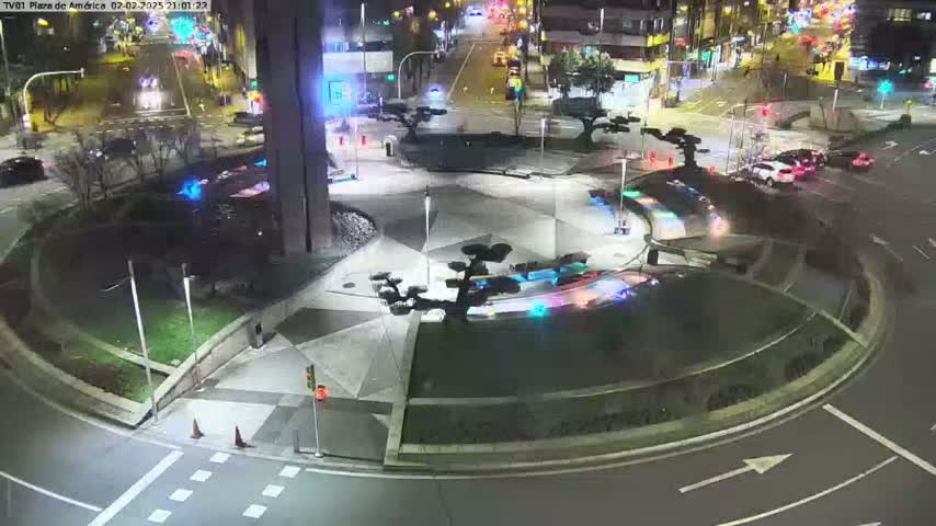 A nighttime aerial view shows a circular plaza with several small trees and lights, surrounded by roadways with cars.