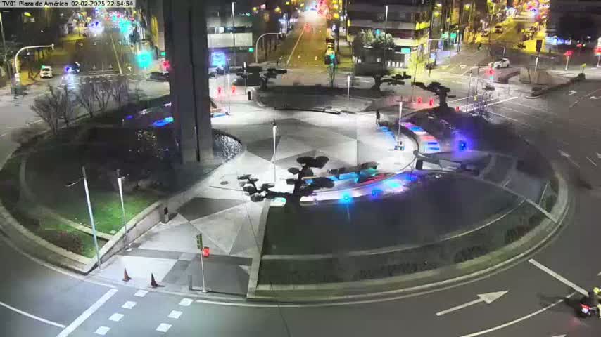 A nighttime aerial view of a roundabout with several vehicles and a few pedestrians, surrounded by landscaping and buildings under clear skies.