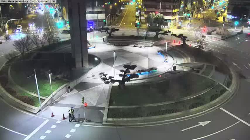A nighttime overhead view of a roundabout with a person on a scooter near the curb,  surrounded by landscaped areas and illuminated by streetlights.