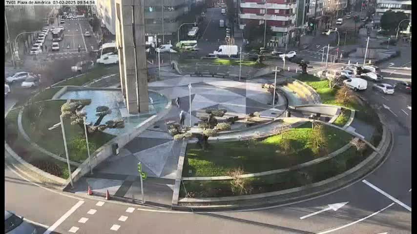A circular plaza with a central fountain and landscaping is surrounded by a roadway with cars, under a sunny sky.
