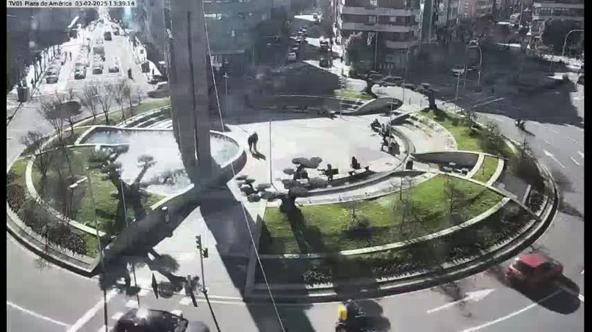A sunny day overlooks a circular plaza with landscaping, benches, and a few pedestrians, surrounded by roads with cars.
