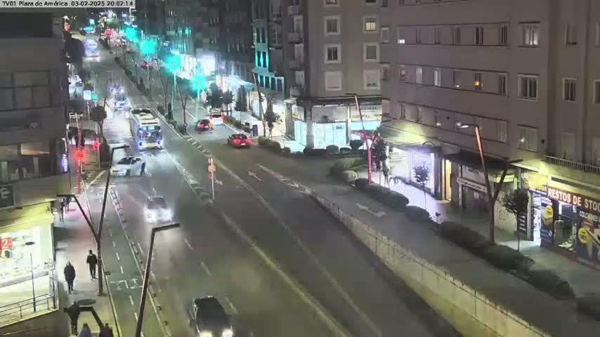 A nighttime, high-angle view of a city street with cars driving on a multi-lane road, pedestrians walking on sidewalks, and buildings lining the street.