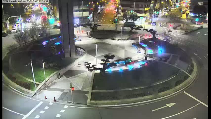 A nighttime aerial view of a circular plaza with illuminated water features and small trees, surrounded by roads with minimal traffic.