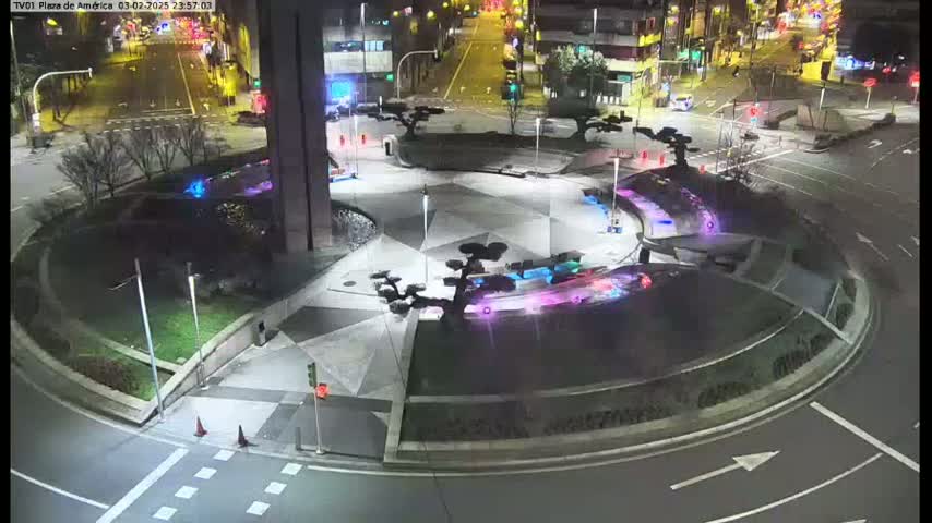 An illuminated plaza at night with geometric patterned pavement, several small trees, and a circular roadway.