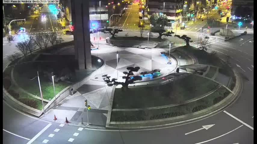 A nighttime aerial view of a circular plaza with geometric landscaping, artificial trees, and a small water feature, surrounded by roads with minimal traffic.