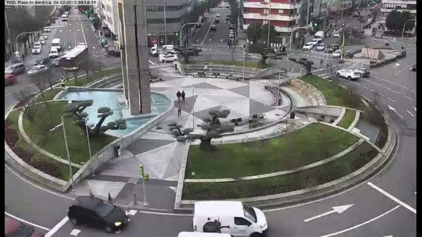 An aerial view of a roundabout with landscaping features, including several small trees and a water feature, with cars and a van driving around it on an overcast day.