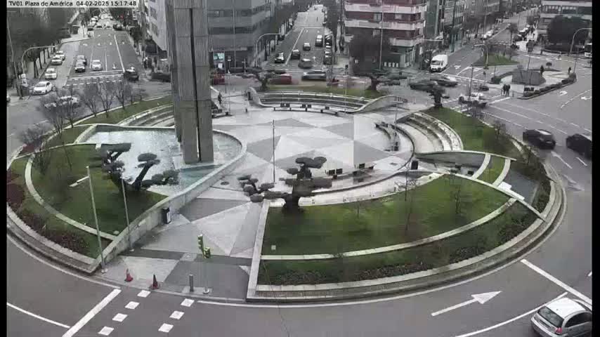 A circular plaza with landscaping features, a central fountain, and surrounding roads with cars is seen on a cloudy day.