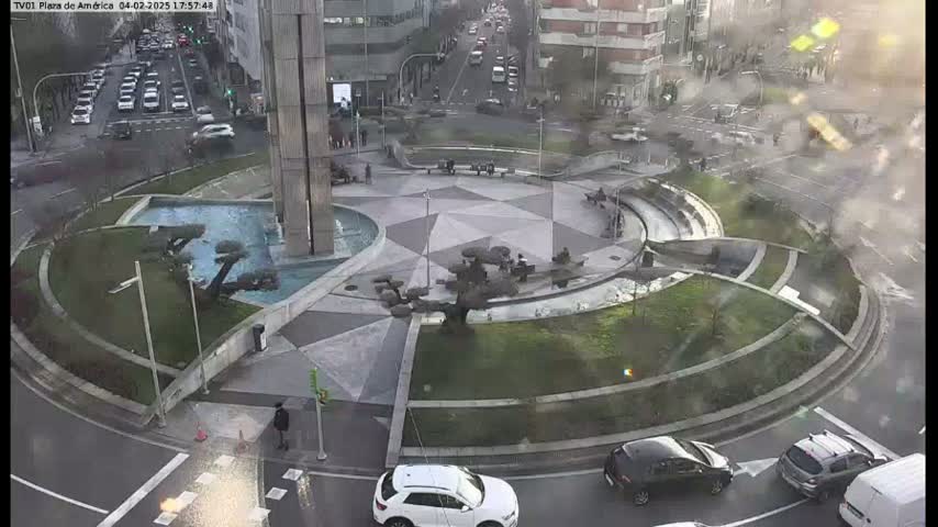 An aerial view of a roundabout with a central fountain and geometric patterned paving, surrounded by cars and pedestrians on a partly sunny day.