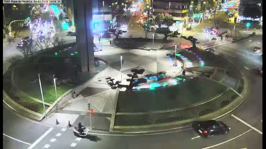 A nighttime aerial view of a roundabout with several vehicles, landscaping, and streetlights.
