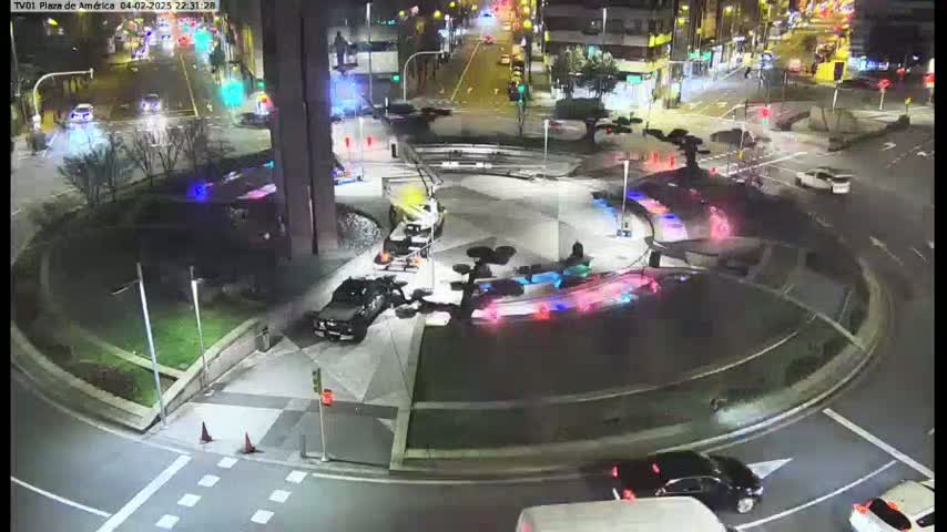 A nighttime aerial view of a roundabout with several vehicles, including police cars, and illuminated features in a plaza.