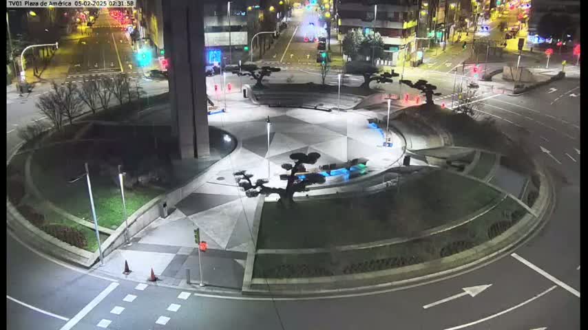 A nighttime aerial view shows a circular plaza with geometric landscaping, small trees, and blue lighting features, surrounded by a roadway.