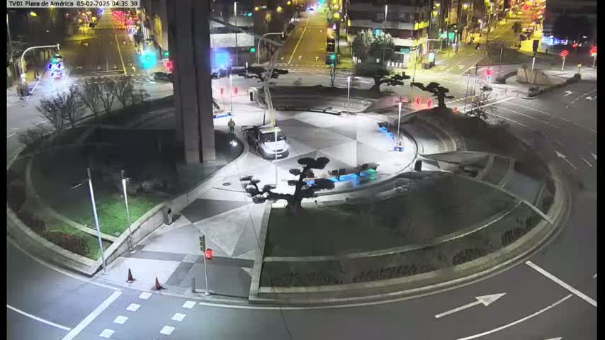 A nighttime aerial view of a circular plaza with landscaping, a small truck, and surrounding roads with traffic.